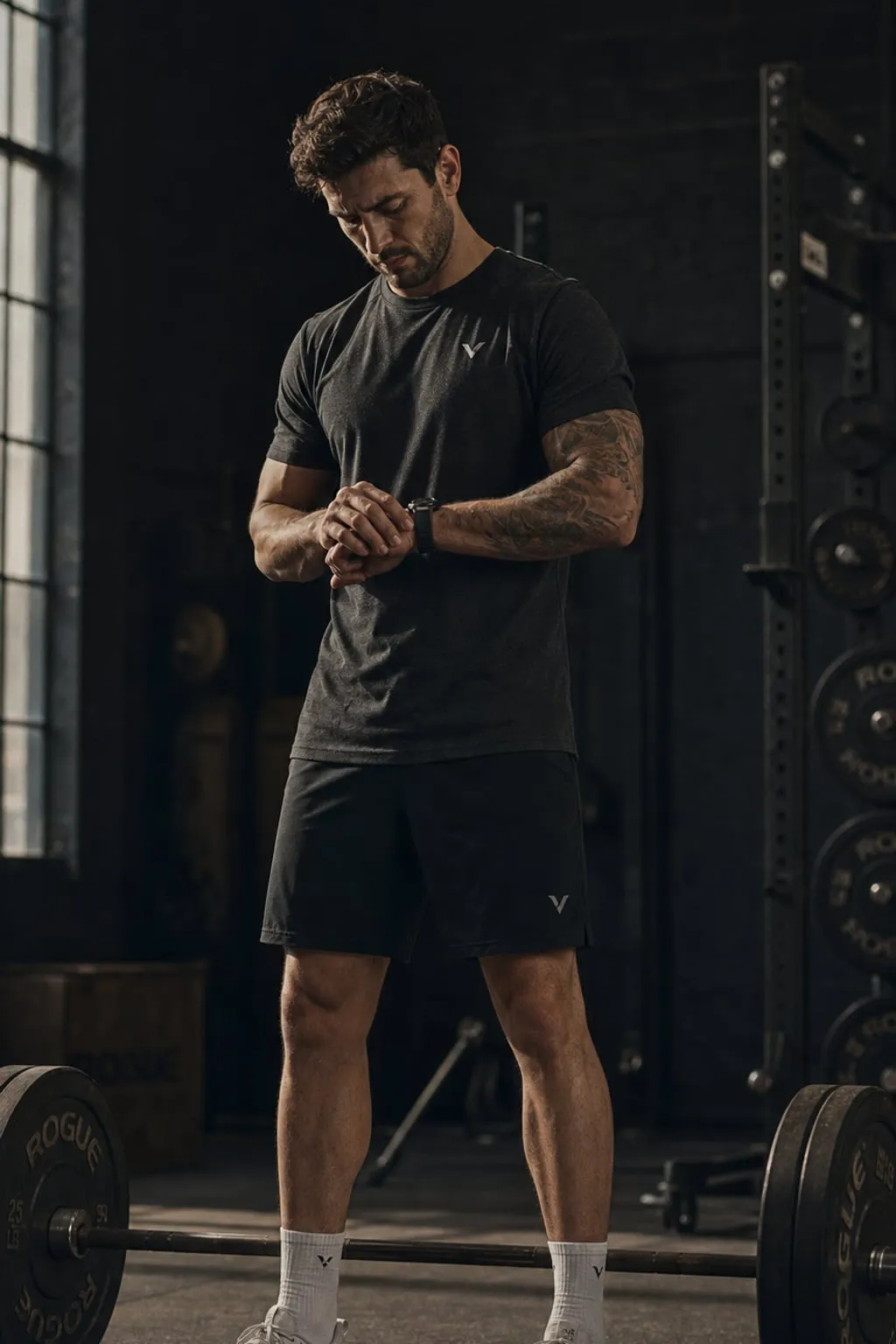 Athlete in a dark gym at dawn checking his wristwatch between sets, a loaded barbell at his feet